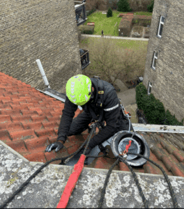 Professionel håndværker i klatreudstyr udfører tætning af skotrende på tag med tagsten ved hjælp af reb og sikkerhedsudstyr i etagebyggeri.